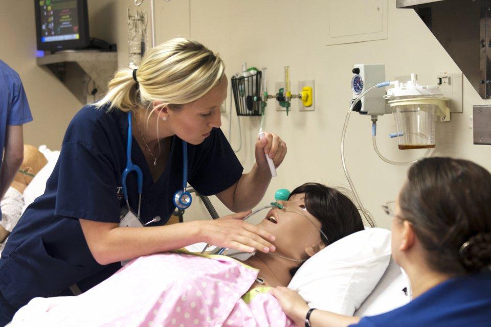 Nursing students in a lab simulation look over a mannequin in a hospital bed.